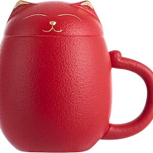 May include: A red ceramic mug shaped like a cat, with a matching lid. The mug has a textured surface and a handle. The cat's face is detailed with gold accents on the ears and nose. The mug is designed for beverages.