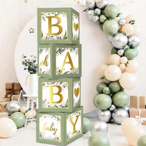 May include: Four green square blocks stacked to spell "BABY" with gold lettering. Each block has a floral design and a gold border. The blocks are decorated with a heart and the words "oh" and "baby". A balloon arch and other decorations are in the background.