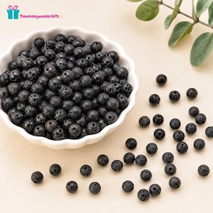 May include: A collection of round, black lava rock beads with a porous, textured surface. Many beads are contained within a white, scalloped-edge bowl, while several individual beads are scattered outside the bowl on a light surface.