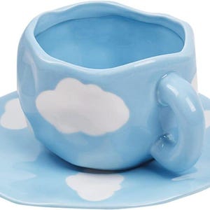 May include: A light blue ceramic teacup and saucer set. The cup and saucer are decorated with white cloud shapes. The cup has a rounded square shape and a curved handle. The saucer is a rounded square shape.