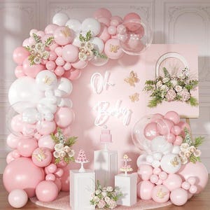 May include: A balloon arch in shades of pink and white featuring clear balloons, gold butterfly accents, and floral arrangements. A pink sign with the white neon text Oh Baby is centered above three white pedestals holding small cakes.