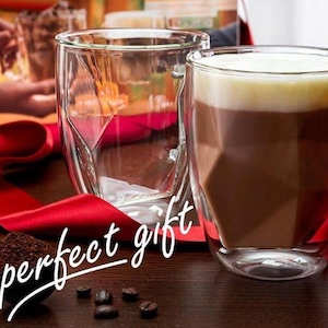 May include: Two clear, double-walled glass mugs, one filled with a layered coffee drink. The mugs have a geometric design. A red ribbon and coffee beans are on the wooden surface. Text reads "The perfect gift."