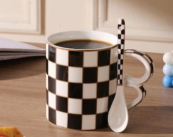 May include: A ceramic coffee mug with a black and white checkered pattern, filled with dark coffee. A matching spoon rests beside the mug. The mug has a gold trim and a unique handle design. The spoon also has a checkered pattern.