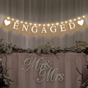 May include: A burlap banner with the word "ENGAGED" in white letters, strung with fairy lights and heart-shaped accents. The banner is displayed above a table decorated with flowers and a "Mr & Mrs" sign, creating a festive atmosphere.