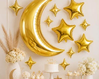 May include: A large gold crescent moon balloon and several gold star balloons of varying sizes arranged on a wall. Below, a white cake on a pedestal, white floral arrangements in vases, and small cupcakes are displayed on a table.