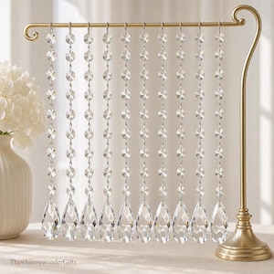 May include: A gold-toned metal stand featuring a horizontal bar from which ten vertical strands of clear, faceted crystal beads hang. Each strand terminates in a large, teardrop-shaped crystal pendant. The text ThewhimsywonderGifts is visible.