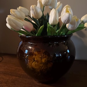May include: A dark brown ceramic vase with a glossy finish, filled with white and pale pink tulips and green leaves. The vase has a decorative yellow and brown floral design. The arrangement sits on a wooden surface.