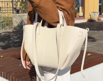 May include: Cream-colored tote bag with long shoulder straps and short handles. The bag has a structured design with a subtle texture and a circular detail on the side. The bag is held in front of a person wearing a brown jacket and white pants.