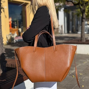 May include: A large, brown leather tote bag with two handles and a shoulder strap. The bag has a simple, classic design with a spacious interior. The bag is carried by a person wearing a black jacket and white pants.
