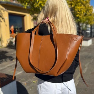 May include: A large, brown leather tote bag with long handles and a shoulder strap. The bag has a simple, elegant design with a button detail on the side. The bag is carried by a person with long blonde hair.