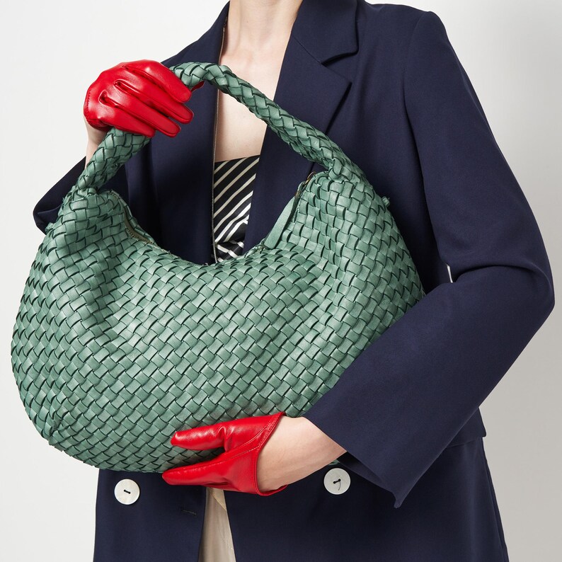 May include: A woven, sage green handbag with a curved shape and braided handle. The bag is held by a person wearing red leather gloves and a navy blue blazer. The blazer has white buttons.