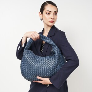 May include: A woven, blue leather handbag with a rounded shape and a braided handle. The bag is held by a person wearing a navy blue blazer. The bag's texture and color suggest a high-end, fashionable accessory.