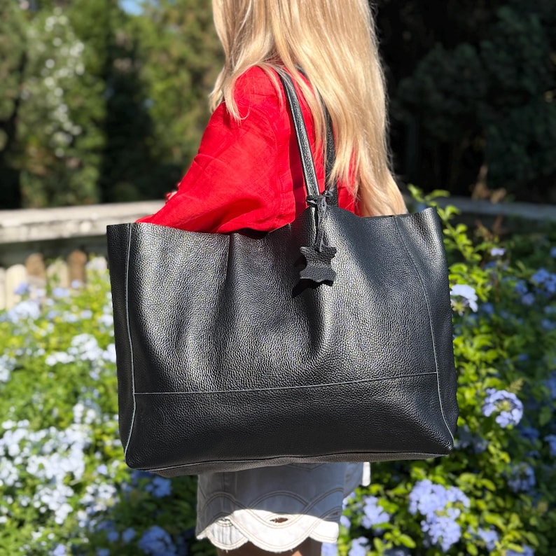 May include: A large black leather tote bag with a textured finish. The bag has a rectangular shape with a handle and a decorative leather charm. The bag is being held in front of a background of green foliage and blue flowers.