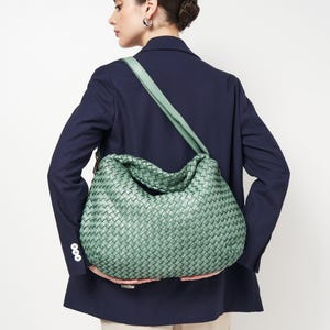 May include: A woven, mint green shoulder bag with a matching strap. The bag has a large, slouchy design and is held by a person wearing a navy blue blazer. The bag's woven pattern is visible.