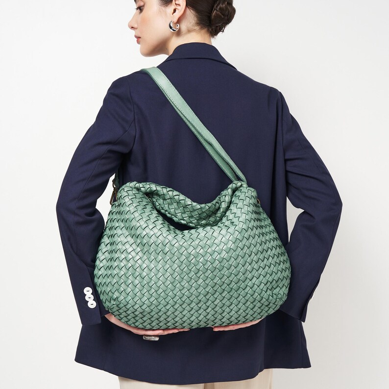 May include: A woven, mint green shoulder bag with a matching strap. The bag has a large, slouchy design and is held by a person wearing a navy blue blazer. The bag's woven pattern is visible.