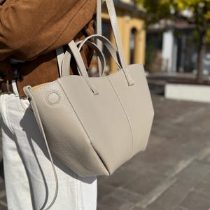 Może przedstawiać: Jasnoszara skórzana torba typu tote o strukturalnym designie. Torba posiada górny uchwyt i pasek na ramię. Torba ma okrągły detal z boku i widoczne szwy. Torba jest trzymana przez osobę ubraną w brązową kurtkę i białe spodnie.