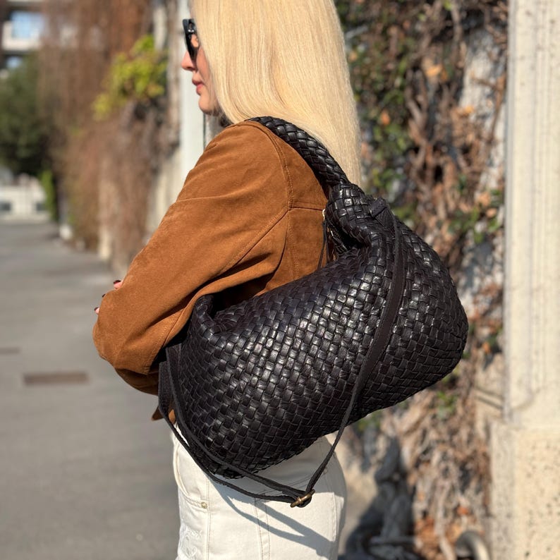 May include: A large, black woven leather shoulder bag with a knotted handle. The bag has a long, adjustable strap and a soft, slouchy shape. The leather appears to be high-quality and the weave is intricate.