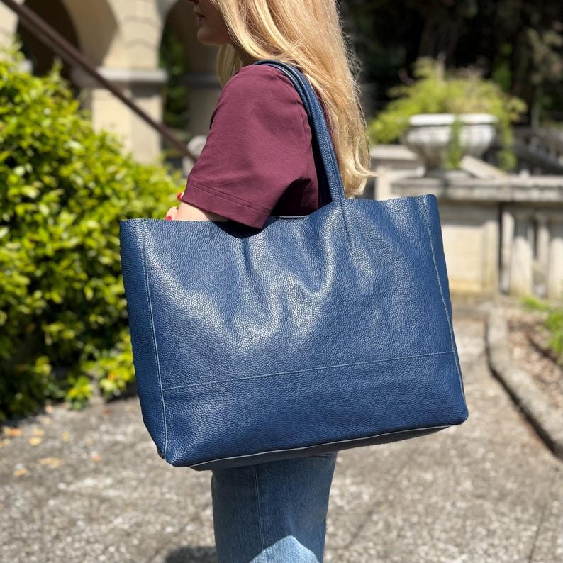 May include: A large, navy blue tote bag with a textured leather appearance. The bag has a rectangular shape with a flat bottom and two handles. The bag is being carried by a person wearing a burgundy shirt and blue jeans.