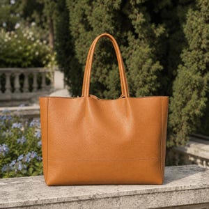 May include: A large, tan leather tote bag with two long handles. The bag has a rectangular shape with a structured design and a subtle texture. The background features greenery and a stone wall.