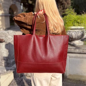 May include: A large, burgundy leather tote bag with two handles. The bag has a rectangular shape with a horizontal seam near the bottom. The leather has a textured appearance. The bag is being held by a person.