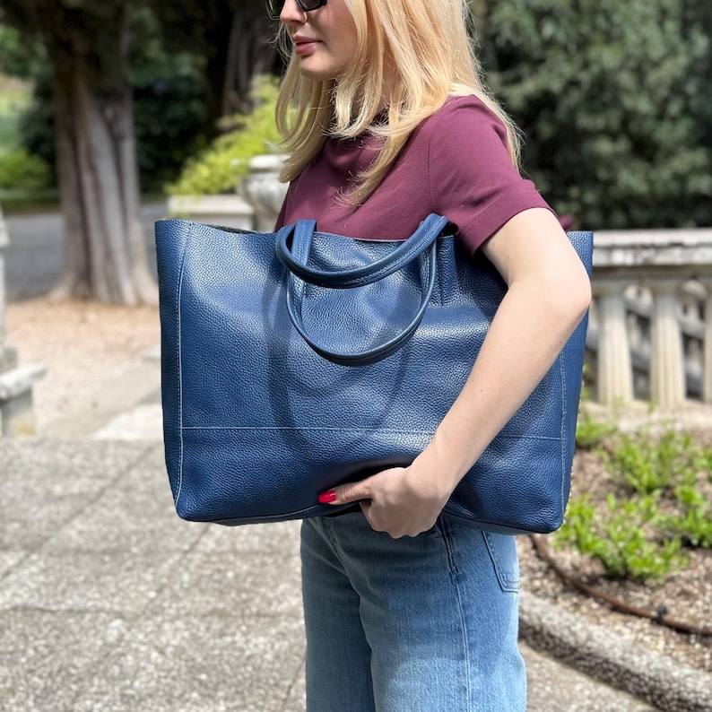 May include: A large, navy blue tote bag with two handles. The bag has a textured surface and is being held by a person wearing a burgundy shirt and blue jeans. The bag appears to be made of leather.