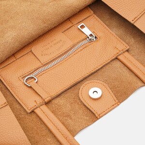 May include: Close-up of a tan leather tote bag interior. The bag features a zippered pocket with a silver zipper and a magnetic snap closure. The interior is lined with a lighter shade of leather.
