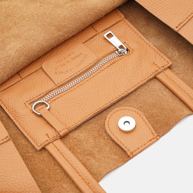 May include: Close-up of a tan leather tote bag interior. The bag features a zippered pocket with a silver zipper and a magnetic snap closure. The interior is lined with a lighter shade of leather.