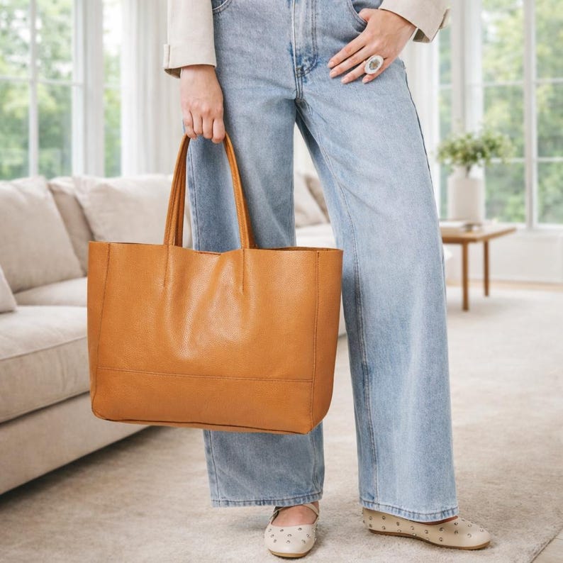 May include: A tan leather tote bag with two handles. The bag is rectangular with a simple design. The person is wearing light blue jeans and tan shoes. The background includes a couch, a table, and a window.