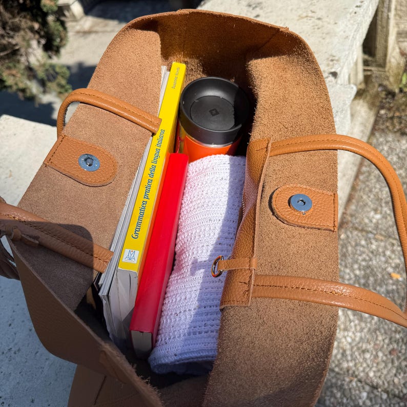 May include: A brown leather tote bag with a light brown trim, containing a yellow book with Italian text, a red book, a white crocheted item, and an orange travel mug. The bag is open, revealing its contents.