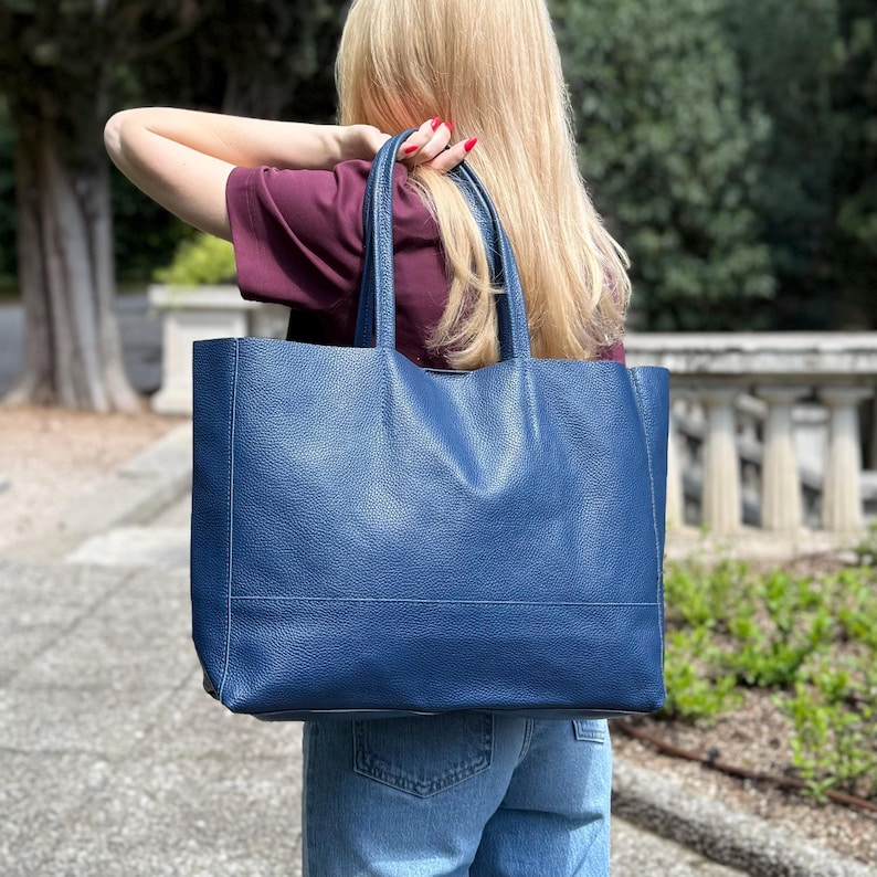 May include: A large, navy blue tote bag with a textured leather appearance. The bag has two handles and a rectangular shape. The bag is being carried outdoors. The background includes greenery and a stone structure.