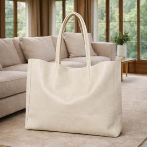 May include: Cream-colored tote bag with two handles. The bag has a textured surface and a rectangular shape. The bag is sitting on a light-colored rug in a living room setting.