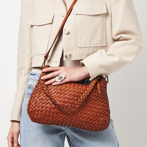 May include: A woven leather shoulder bag in a warm brown hue. The bag features a braided strap and a long, adjustable shoulder strap. The bag is worn across the body. The bag is a medium size.