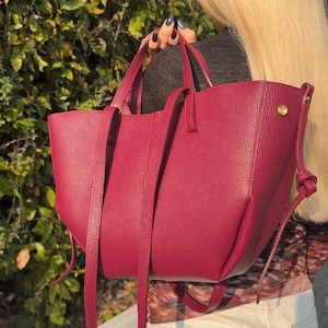 May include: A burgundy leather tote bag with a structured design. The bag features two top handles and a long, adjustable shoulder strap. A gold-colored metal rivet is visible on the side. The bag is held against a background of green foliage.