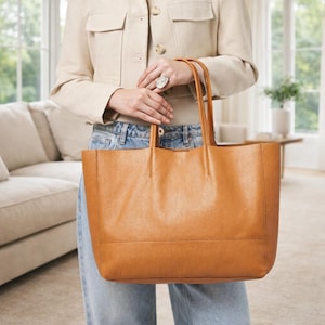 May include: A large, tan leather tote bag with two handles. The bag has a rectangular shape and a textured surface. The bag is being held by a person wearing a tan jacket and blue jeans. The background includes a couch and a window.