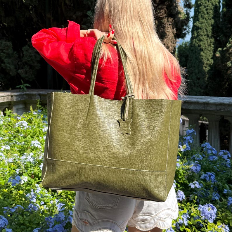 May include: Olive green leather tote bag with a rectangular shape and white stitching details. The bag has two handles and a small leather tag. The bag is carried over the shoulder. The background features a garden with blue flowers and green trees.