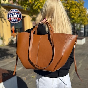 May include: A large, cognac-colored leather tote bag with a pebbled texture, featuring two long shoulder straps, a wide open-top design, and a small circular embossed detail on the upper right side, carried over the shoulder.