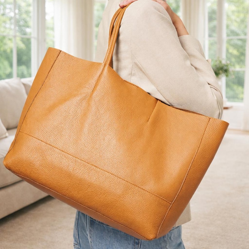 May include: A large, tan leather tote bag with two long handles. The bag has a rectangular shape with a textured surface and visible stitching details. The bag is being held by a person wearing a light-colored blazer and blue jeans.