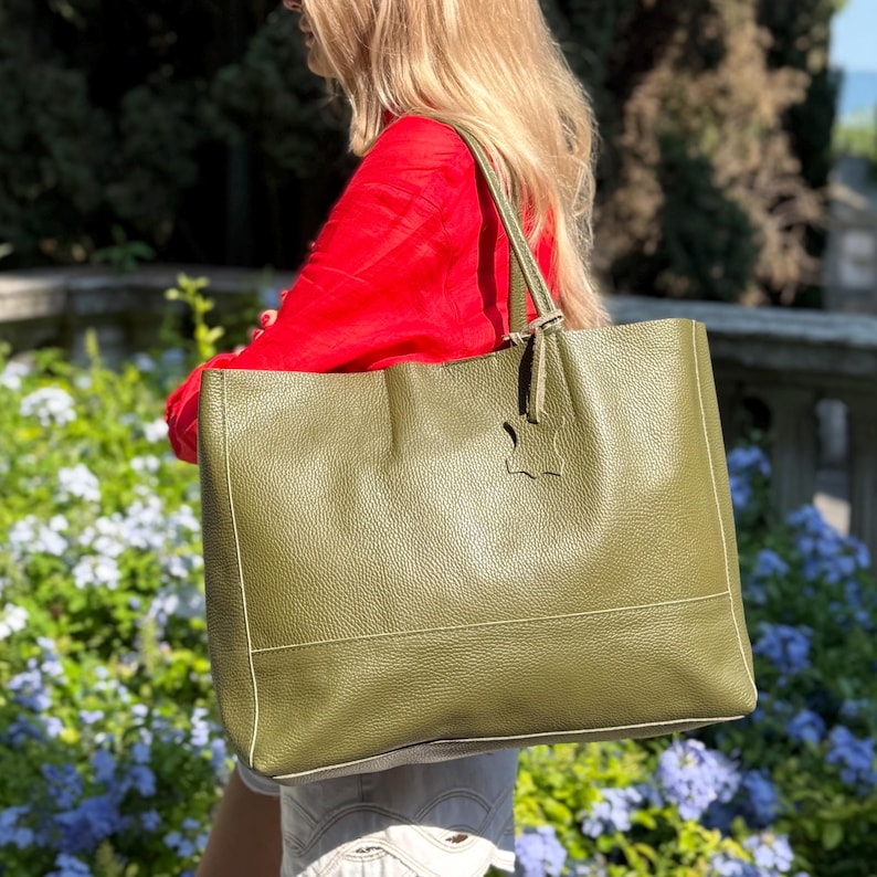 May include: Olive green leather tote bag with a rectangular shape and a top handle. The bag features a textured surface and a contrasting white stitch detail. A small leather tag hangs from the handle. The bag is carried over the shoulder.