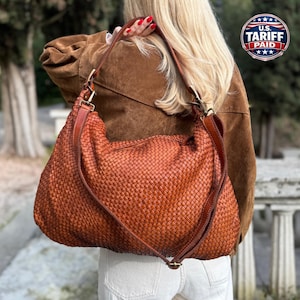 May include: A large, woven brown leather shoulder bag with a long strap. The bag has a textured, basket-weave design and is carried over the shoulder. The bag is a warm, reddish-brown color.