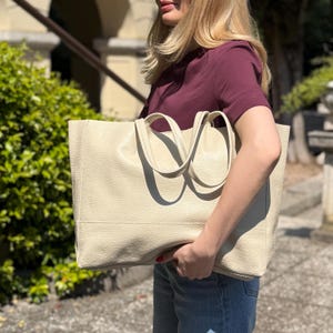 May include: Cream-colored tote bag with two handles. The bag has a rectangular shape and a textured surface. The handles are curved and attached to the top of the bag. The bag is held by a person wearing a burgundy shirt and blue jeans.