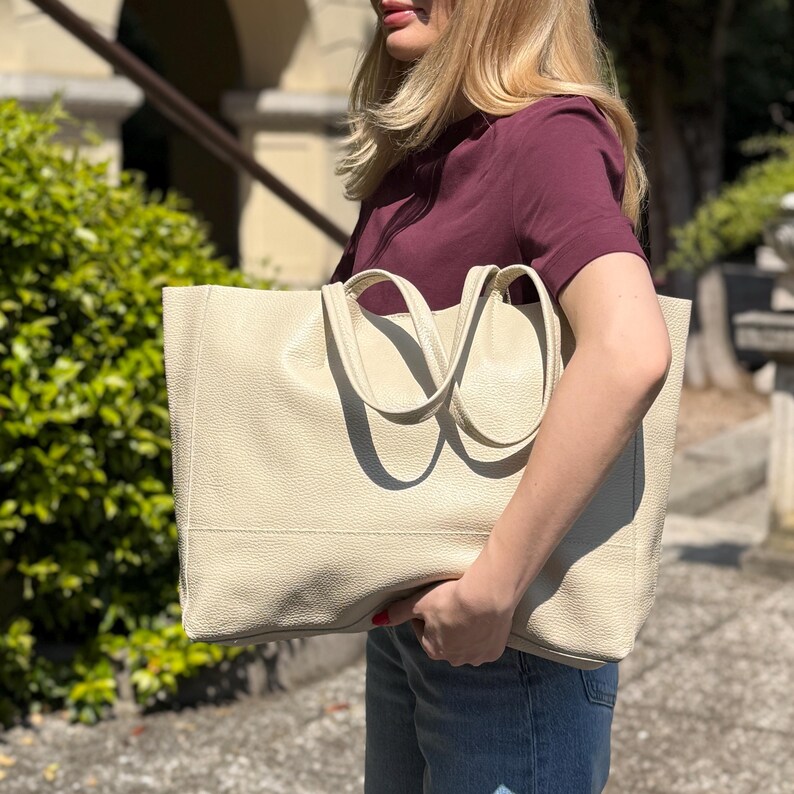 May include: Cream-colored tote bag with two handles. The bag has a rectangular shape and a textured surface. The handles are curved and attached to the top of the bag. The bag is held by a person wearing a burgundy shirt and blue jeans.