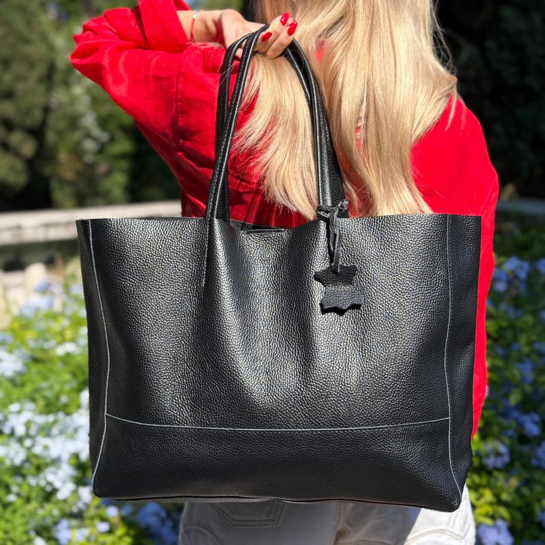 May include: A large black tote bag with a textured leather appearance. The bag has two long handles and a small leather tag attached. The bag is rectangular with a horizontal seam detail. The bag is being held by a person wearing a red shirt.