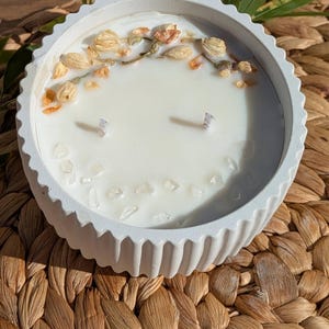 Puede incluir: Una vela blanca y redonda en un cuenco de hormigón texturizado. La vela está decorada con flores secas y cristales. Se ven dos mechas. La vela está sobre una superficie tejida y marrón.