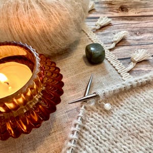 May include: A close-up of a knitting project with a pair of knitting needles and a pearl bead stitch marker. The knitting is a light brown colour and the needles are silver. The project is sitting on a beige fabric surface with a candle and a green stone.