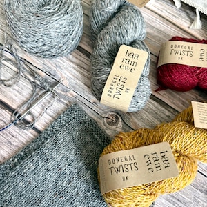 May include: A selection of yarn skeins in shades of grey, burgundy, and yellow, alongside knitting needles and a partially knitted item. The labels on the yarn read "Donegal Twists DK" and "baa ram ewe".