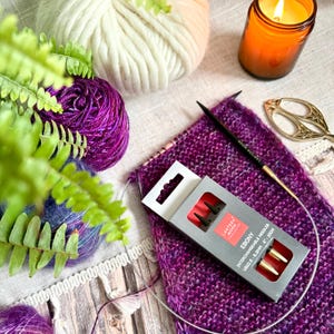Peut inclure: Nature morte avec des fournitures de tricot. Une écharpe violette et rose partiellement tricotée est à côté d'une boîte d'aiguilles à tricoter interchangeables. Il y a des pelotes de laine blanche et violette, une bougie allumée et des ciseaux dorés.