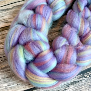 May include: A thick, braided length of roving wool featuring a soft, multi-coloured palette of pastel lavender, light blue, pale yellow, and soft pink. The fibre has a fluffy, unspun texture with visible strands twisted into a continuous braid.