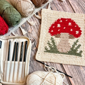 May include: A cream-coloured crochet square with a red mushroom design, green and brown accents. The image includes various coloured yarn balls, a crochet hook, and a black crochet hook set in a beige case.