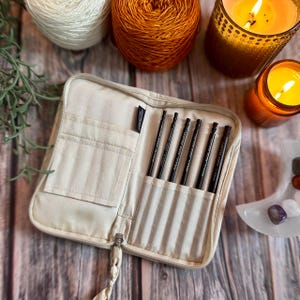 Könnte beinhalten: Eine beige Reißverschlusstasche mit einer kleinen Tasche auf der Vorderseite, geöffnet, um acht schwarze Stricknadeln mit goldener Beschriftung "Knitter's Pride Nova Cubics 3.5mm" zu zeigen.