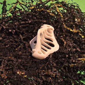Puede incluir: Objeto blanco de pl&aacute;stico impreso en 3D con un dise&ntilde;o esquel&eacute;tico, que se asemeja a una caja tor&aacute;cica, sobre un fondo de papel triturado negro y dorado.